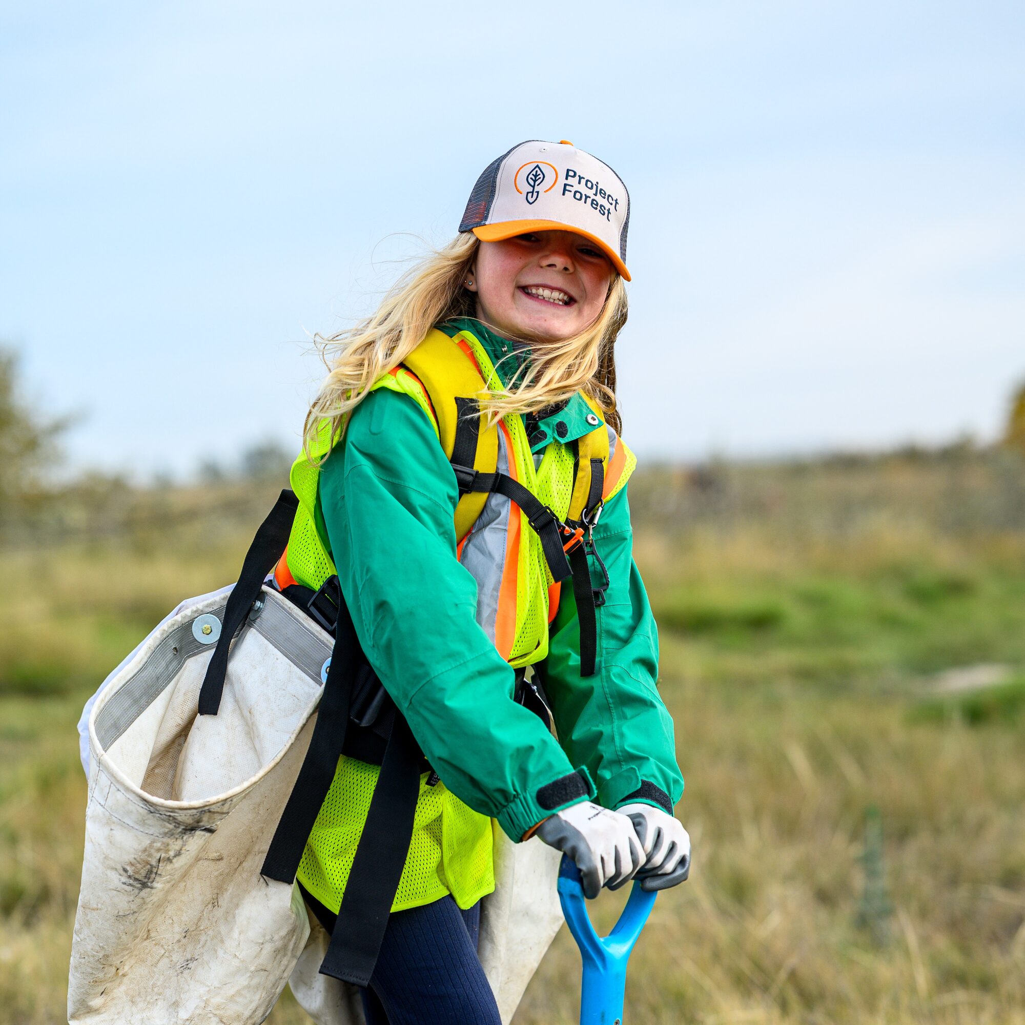 A smiling young Junior Forest Warden in a safety vest holding a tree-planting bag and shovel